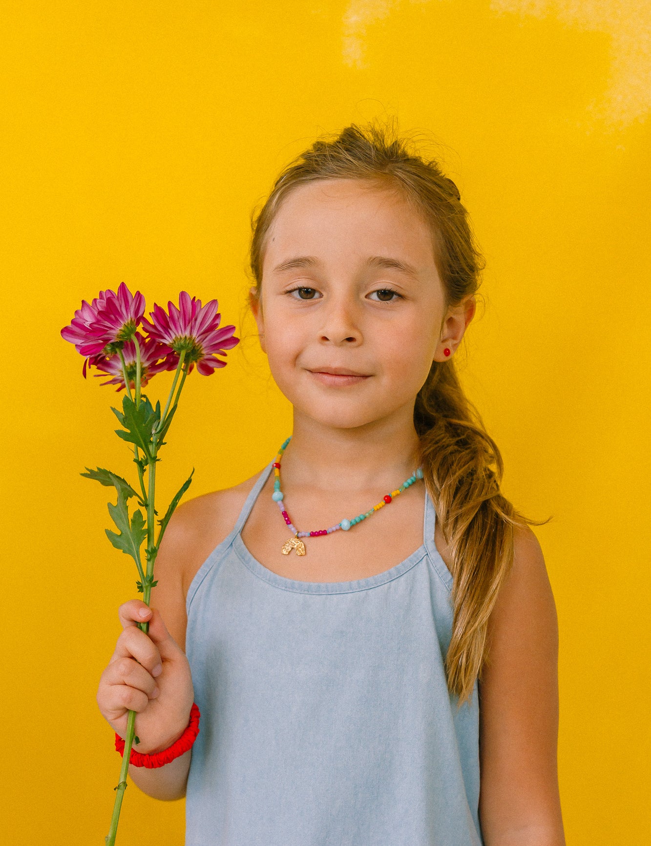 niña con collar arcoíris, sosteniendo  flores sobre fondo amarillo.