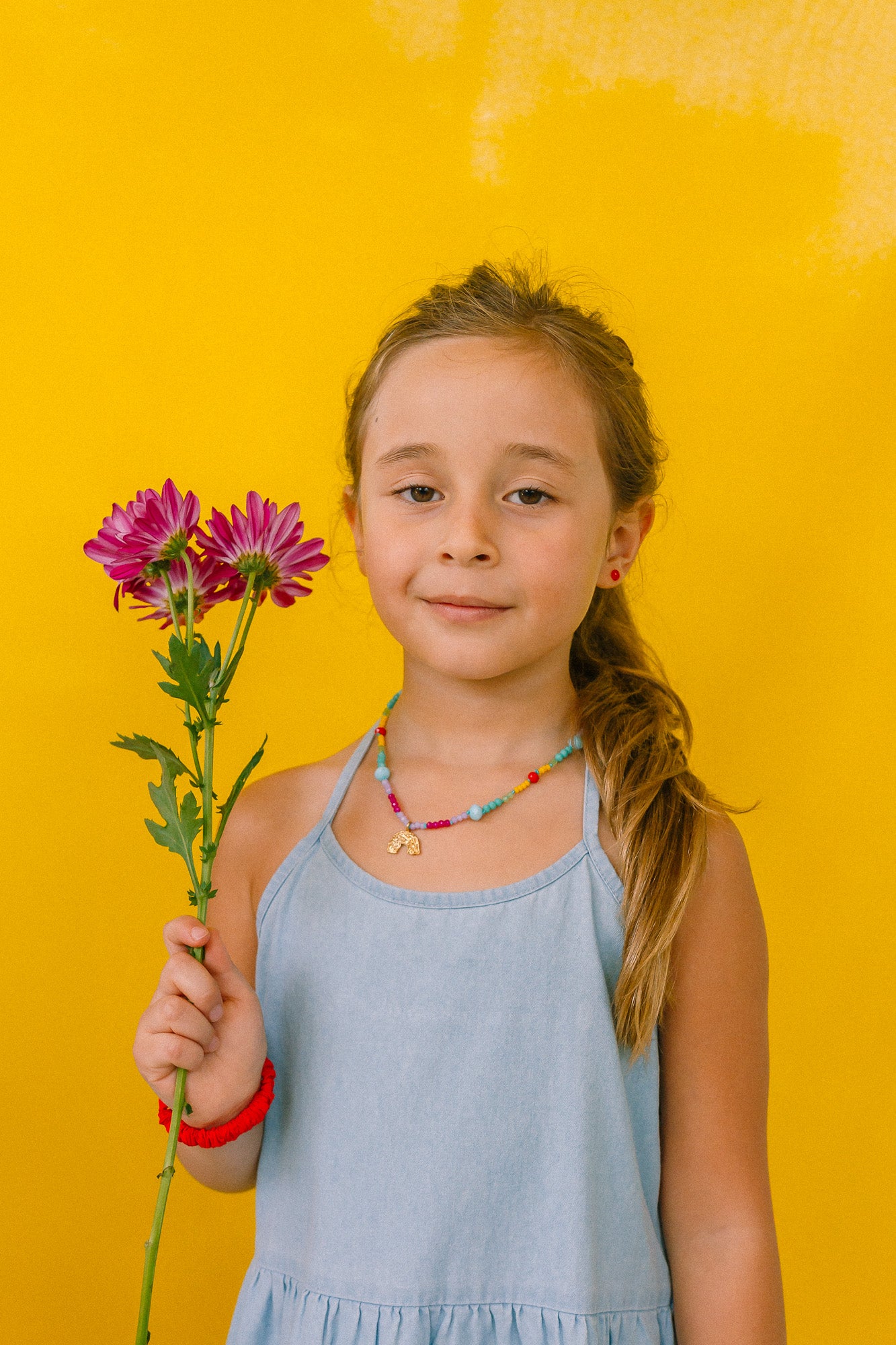 niña con collar arcoíris, sosteniendo flores sobre fondo amarillo.