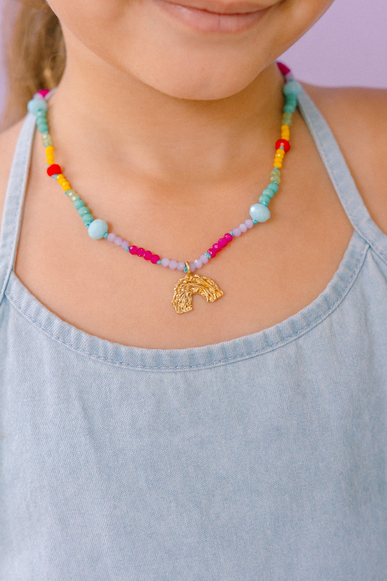 Collar de cuentas multicolor con un colgante dorado en forma de arcoíris, que lleva una niña con un top azul claro.
