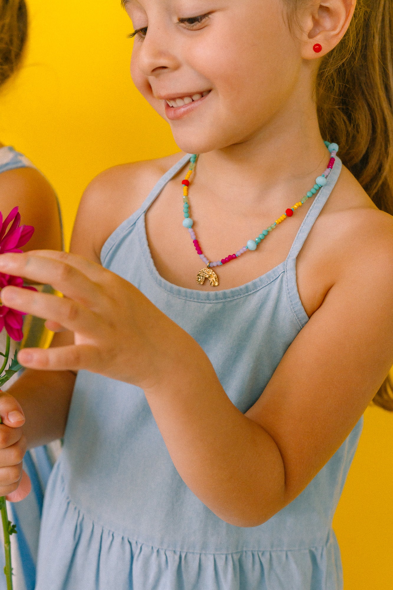 niña de medio perfil usando un collar de colores y sosteniendo una flor rosa sobre un fondo amarillo.