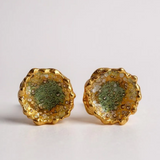 Disposición plana de un par de aretes Topos Coca M en bronce bañado en oro, pintados a mano en verde sage con granulación de vidrio, sobre fondo blanco.
