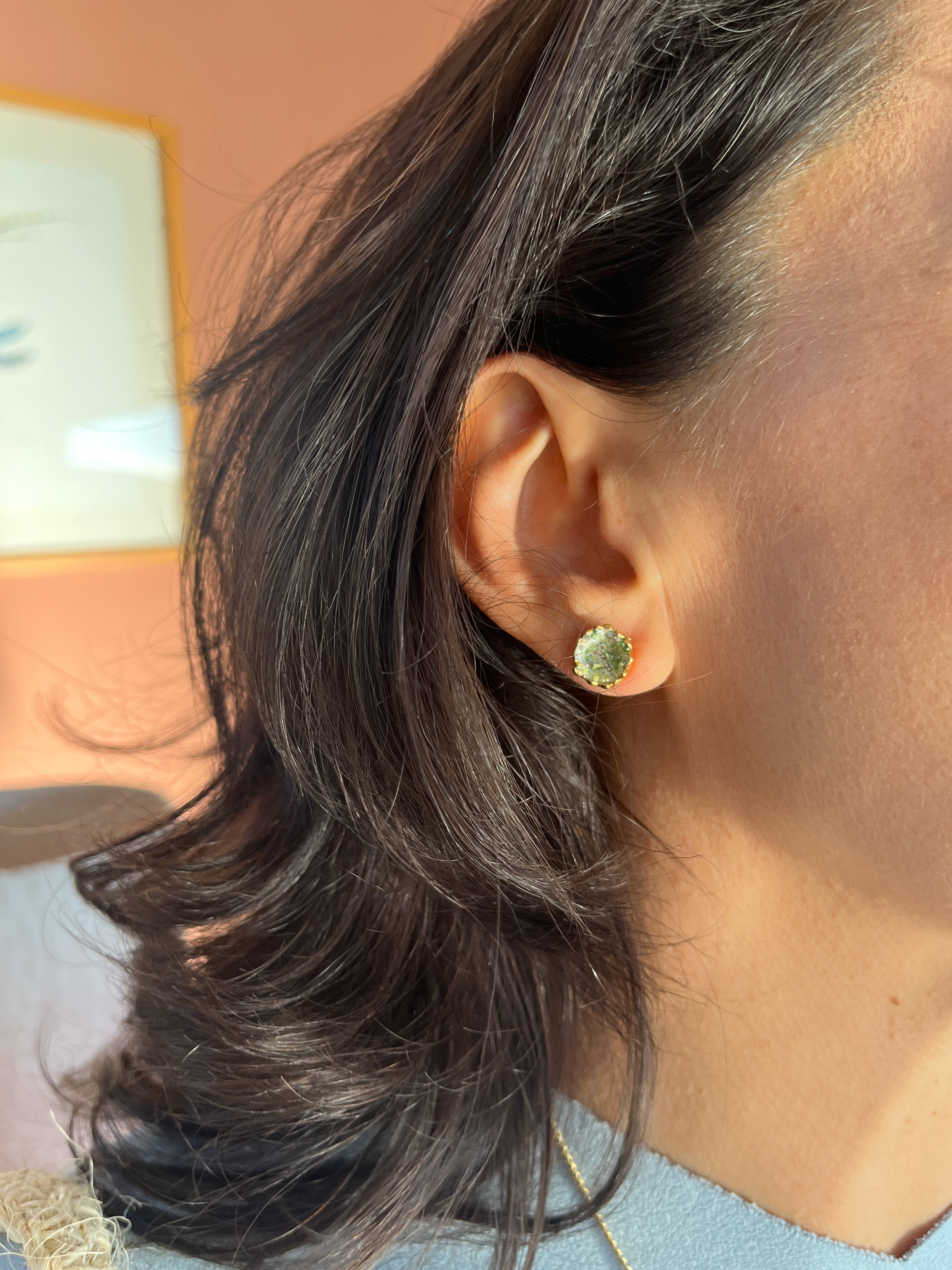Primer plano lateral de la oreja de una mujer con un Topos Coca M en sage pintado a mano con granulación de vidrio en montura de bronce bañado en oro, cabello recogido, sobre fondo neutro.