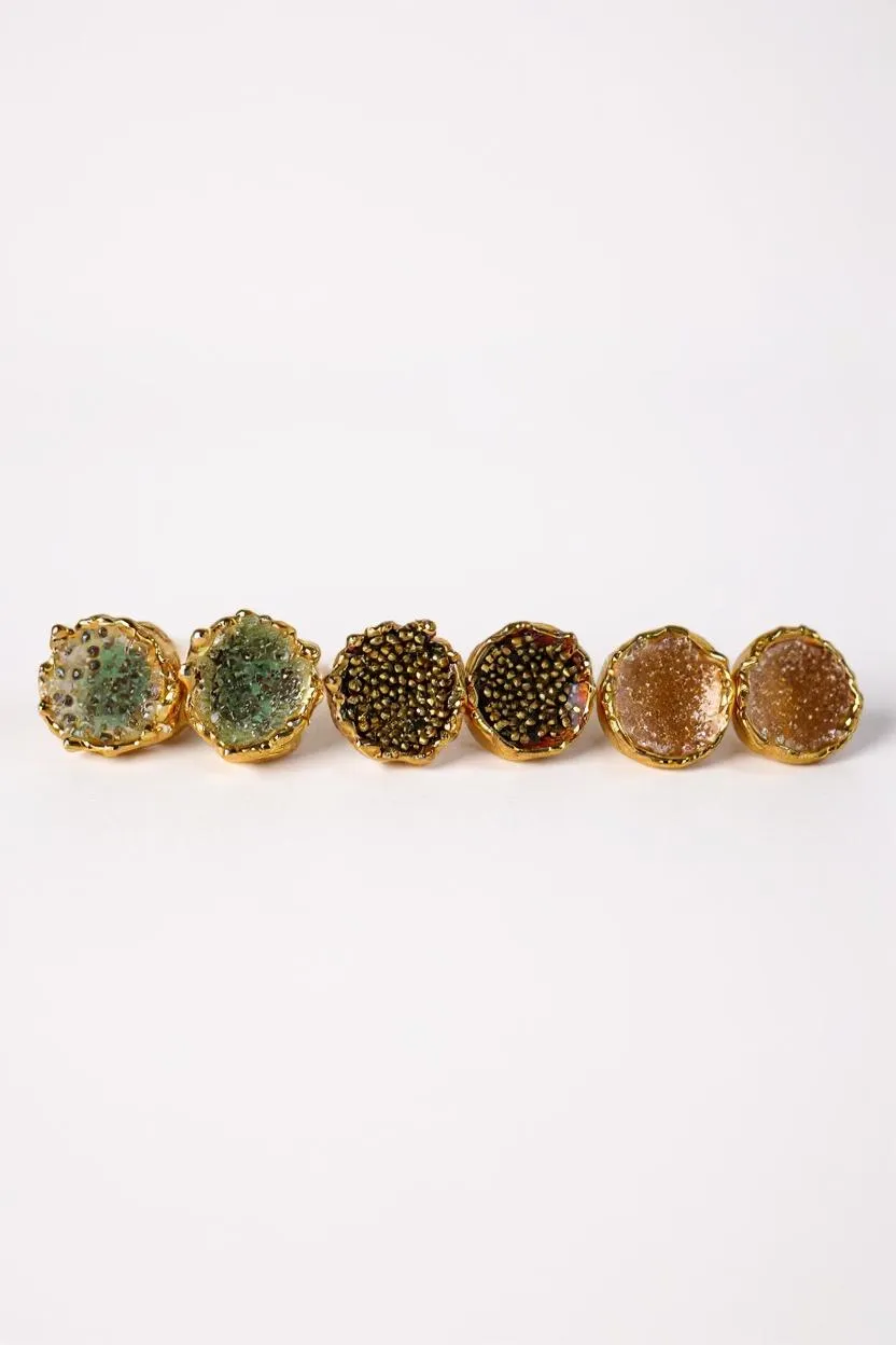 Disposición plana de seis aretes Topos Coca M sobre fondo blanco, mostrando las tres variantes pintadas a mano: dos en verde salvia con granulación de vidrio, dos en rust con granulación de vidrio y dos en tono sunset con granulación turquesa, todas con montura de bronce bañado en oro.