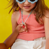 Niña con gafas de sol de montura blanca sostiene el colgante del Collar Fresa cerca de su rostro sobre un fondo amarillo.