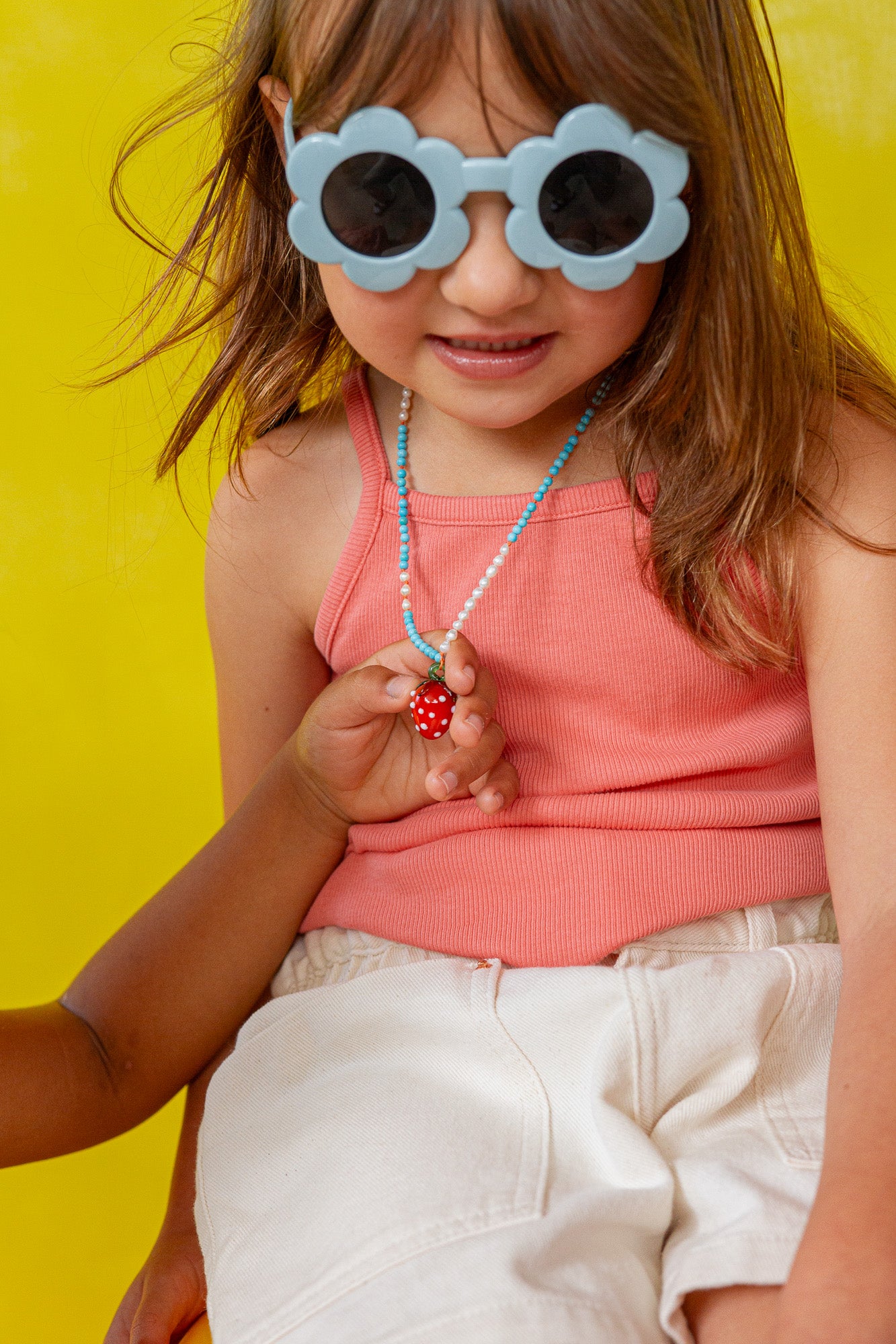 Niña con gafas de sol de montura blanca sostiene el colgante del Collar Fresa cerca de su rostro sobre un fondo amarillo.