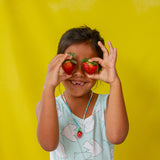 Niña con camiseta sin mangas rosa y pantalones cortos blancos sostiene el colgante del Collar Fresa cerca de su rostro sobre un fondo amarillo