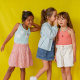 Retrato de cuerpo entero de tres niñas con atuendos veraniegos coloridos y el Collar Fresa, posando de forma divertida frente a un fondo amarillo.