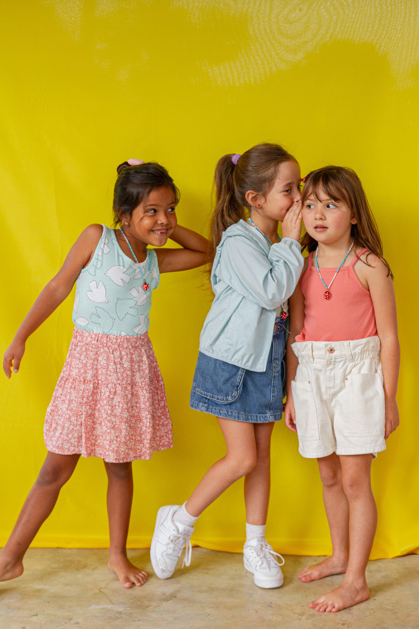 Retrato de cuerpo entero de tres niñas con atuendos veraniegos coloridos y el Collar Fresa, posando de forma divertida frente a un fondo amarillo.