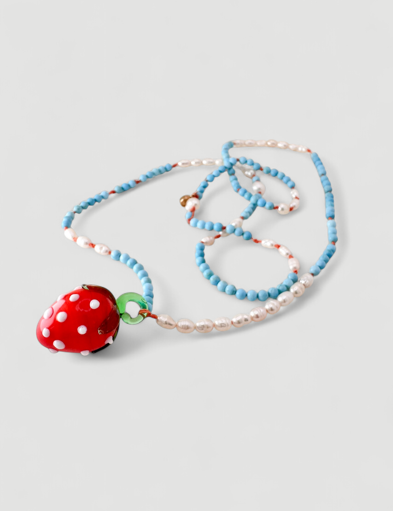 Collar Fresa dispuesto verticalmente sobre fondo blanco, mostrando cuentas alternadas turquesas y blancas y un colgante de fresa de vidrio rojo.