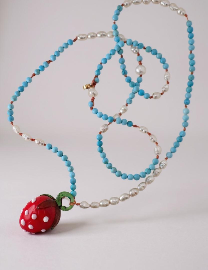 Disposición plana del Collar Fresa sobre fondo blanco, mostrando la cadena de cuentas enrollada ordenadamente con el colgante de fresa de vidrio rojo.