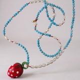 Disposición plana del Collar Fresa sobre fondo blanco, mostrando la cadena de cuentas enrollada ordenadamente con el colgante de fresa de vidrio rojo.