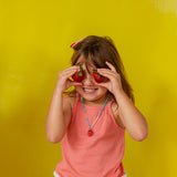 Niña con camiseta sin mangas color coral sostiene el colgante del Collar Fresa frente a su ojo sobre un fondo amarillo brillante.