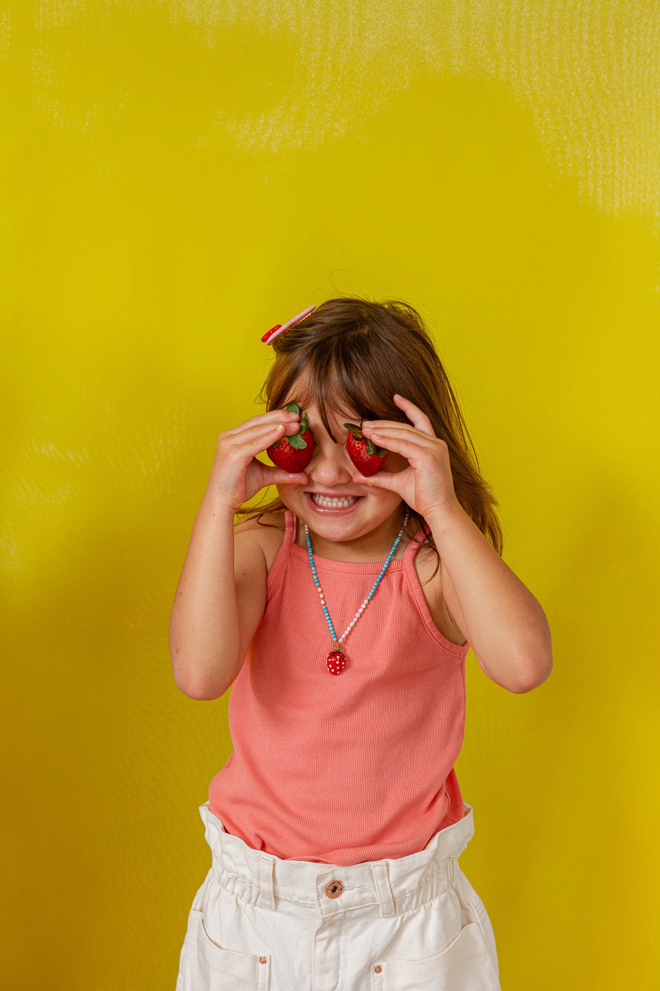 Niña con camiseta sin mangas color coral sostiene el colgante del Collar Fresa frente a su ojo sobre un fondo amarillo brillante.