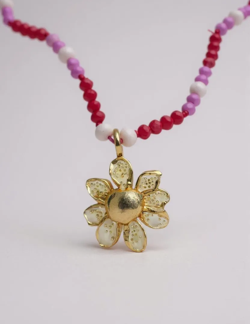 primer plano Collar Margarita en bronce bañado en oro con dije pintado a mano y cristales en tonos rosa y fucsia, sobre fondo blanco.