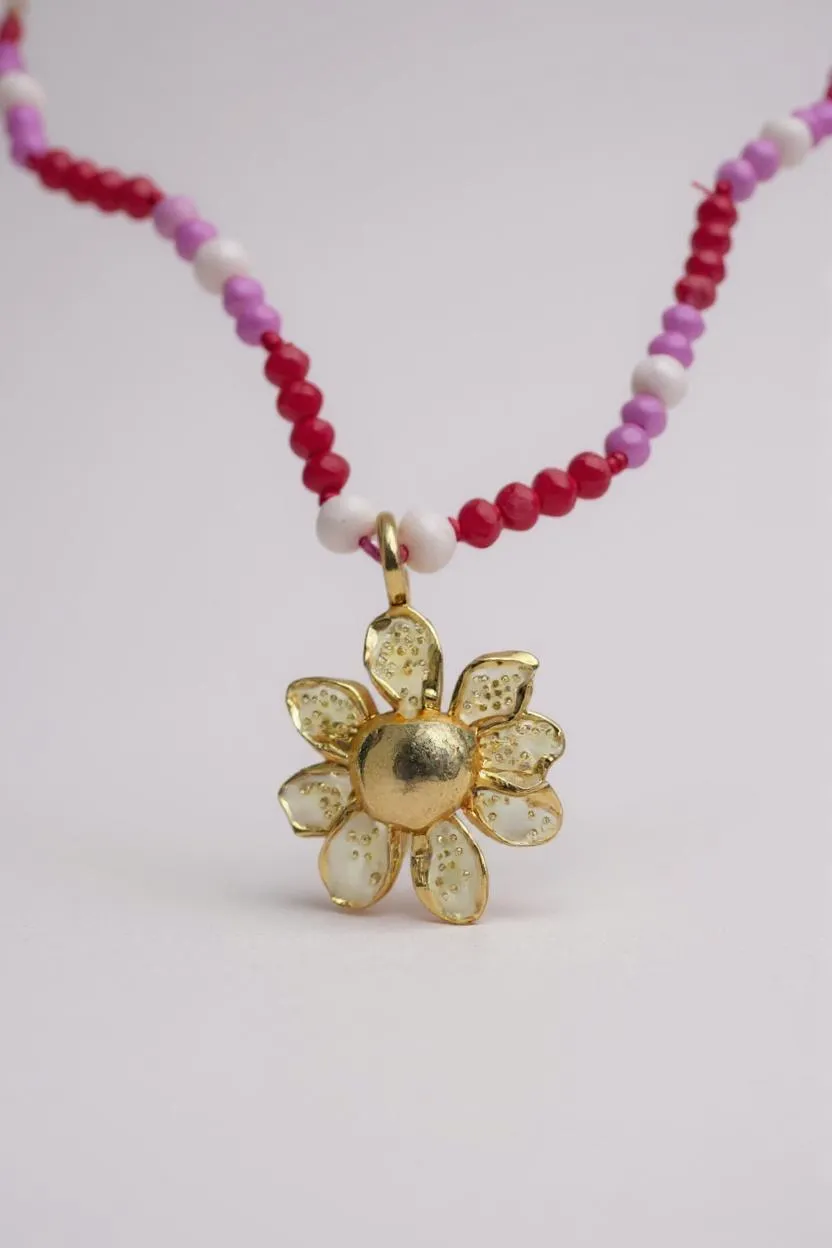 primer plano Collar Margarita en bronce bañado en oro con dije pintado a mano y cristales en tonos rosa y fucsia, sobre fondo blanco.