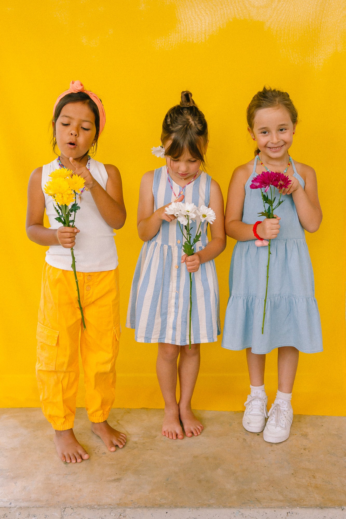 Tres niñas sosteniendo flores sobre un fondo amarillo, usando callares de cuentas multicolor.