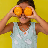 Niña con blusa sin mangas verde menta sostiene dos naranjas sobre los ojos, sonriendo frente a un fondo amarillo brillante.