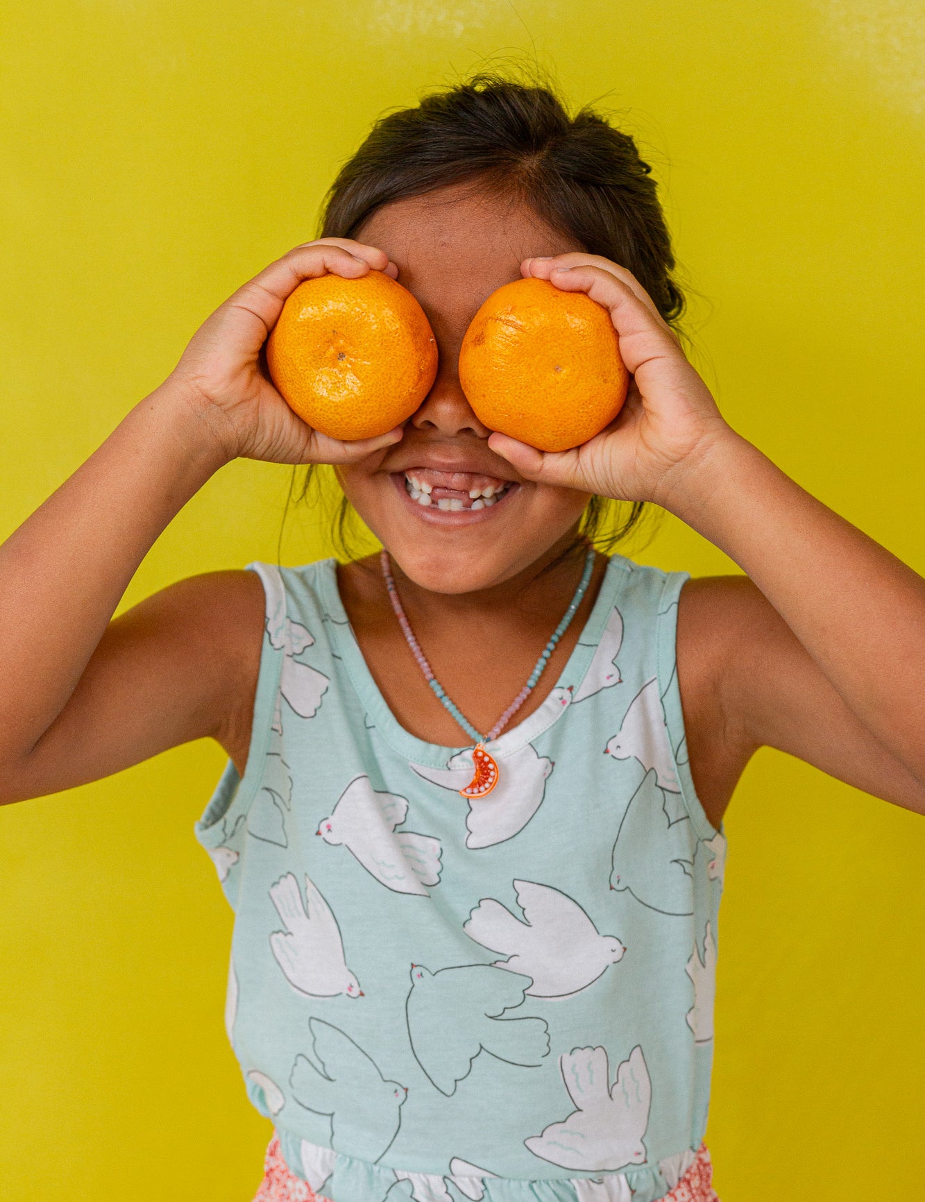 Niña con blusa sin mangas verde menta sostiene dos naranjas sobre los ojos, sonriendo frente a un fondo amarillo brillante.