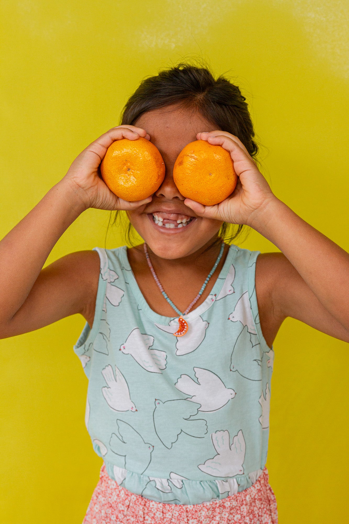 Niña con blusa sin mangas verde menta sostiene dos naranjas sobre los ojos, sonriendo frente a un fondo amarillo brillante.
