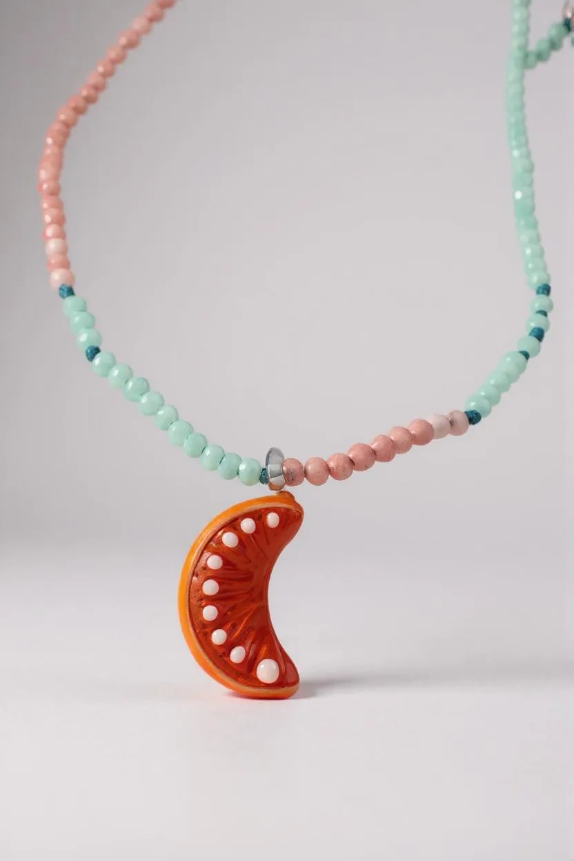 Collar Naranja enrollado sobre fondo blanco, mostrando varias vueltas de cuentas en verde menta y rosa con el colgante de media luna naranja visible.