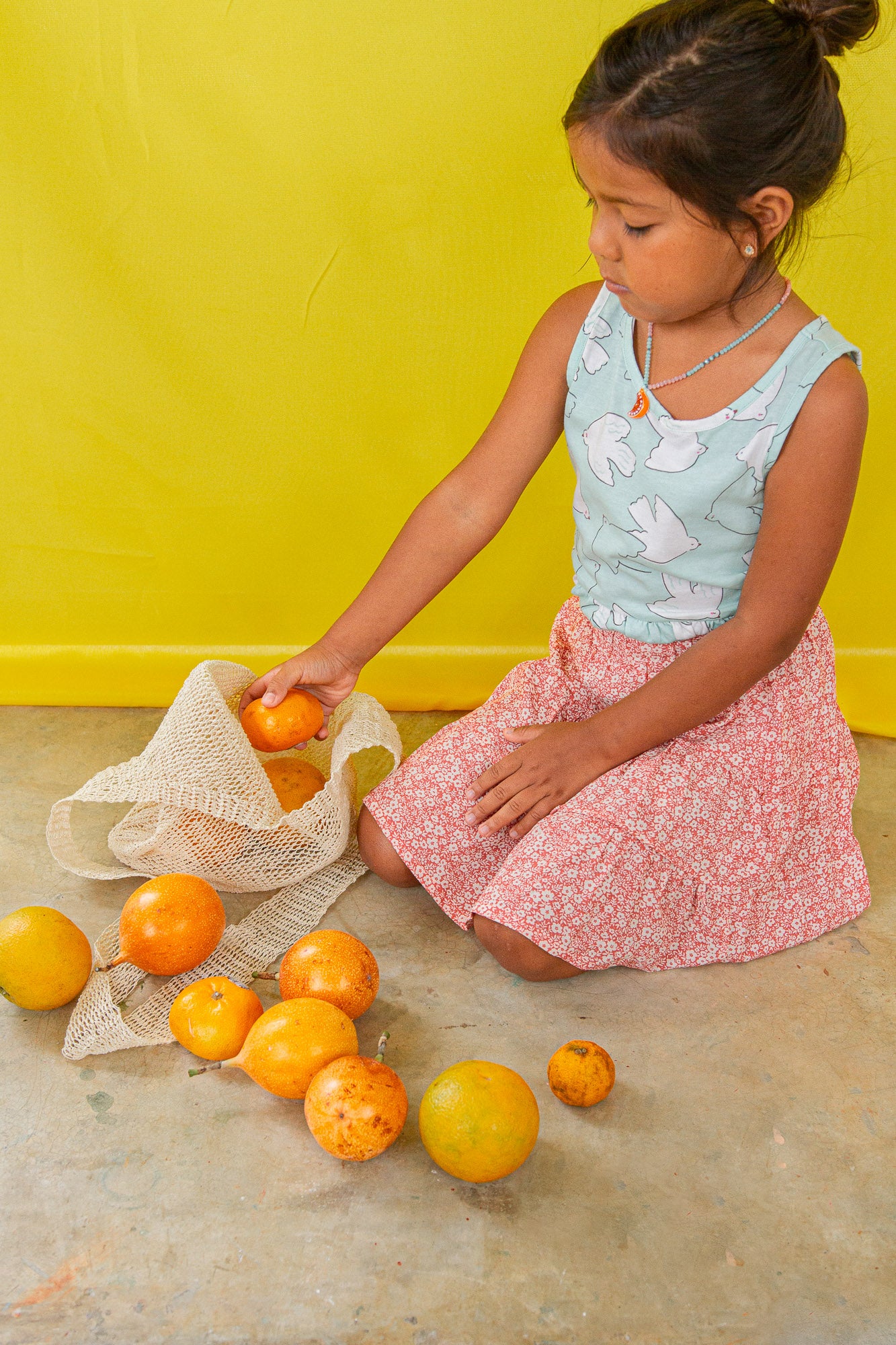Niña arrodillada en el suelo con blusa verde menta y falda pastel juega con naranjas y lleva puesto el Collar Naranja