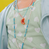 “Niña arrodillada en el suelo con blusa verde menta y falda pastel juega con naranjas y lleva puesto el Collar Naranja.