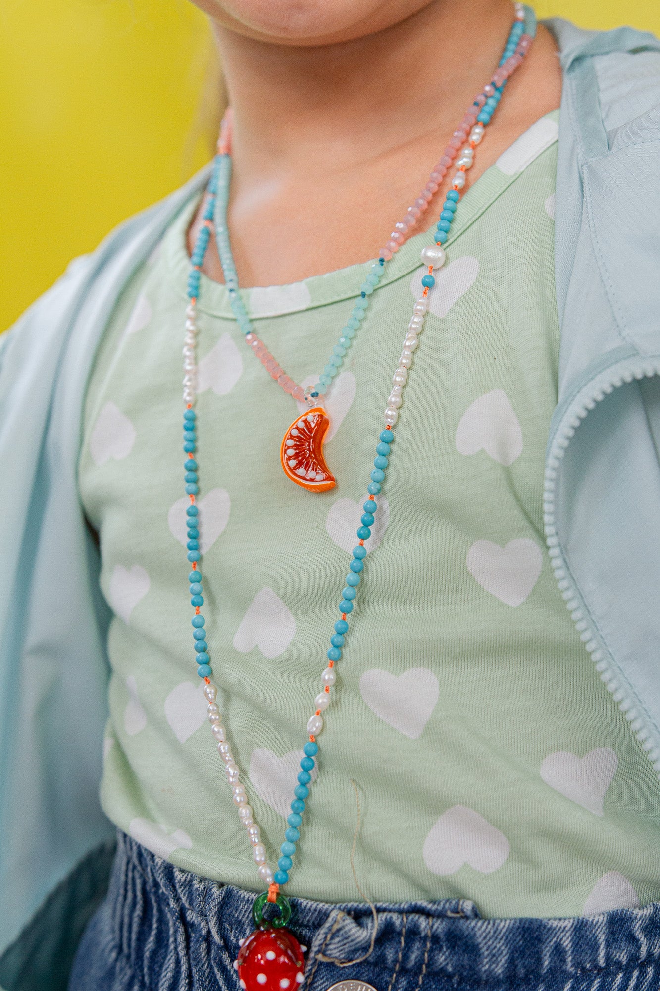 “Niña arrodillada en el suelo con blusa verde menta y falda pastel juega con naranjas y lleva puesto el Collar Naranja.