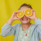 Niña con chaqueta de mezclilla y camiseta verde menta sostiene dos naranjas sobre los ojos frente a un fondo amarillo.