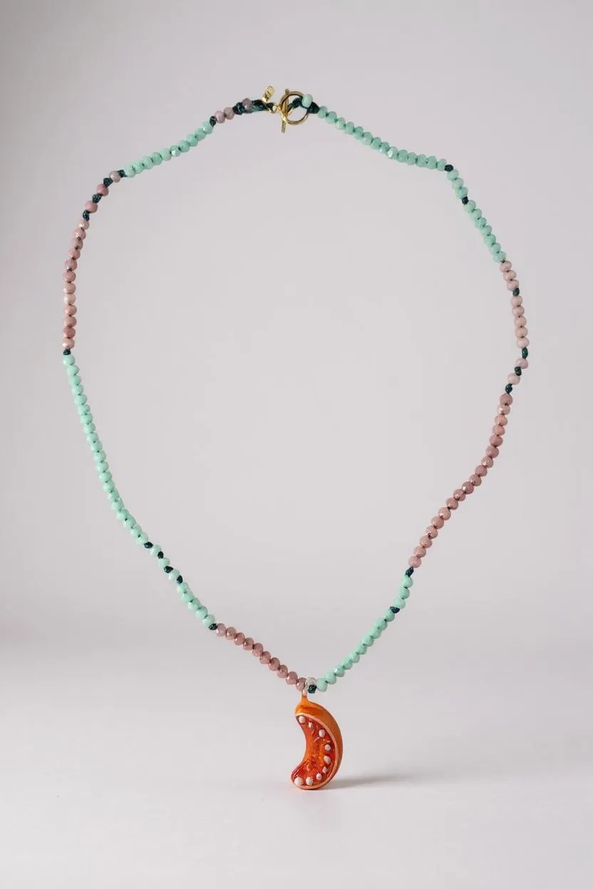 Collar Naranja dispuesto en una suave curva sobre superficie blanca, resaltando las cuentas en verde menta y rosa y el colgante de media luna naranja.
