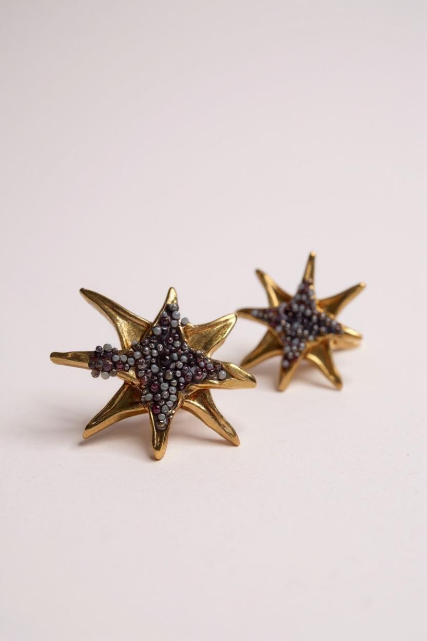 Par de aretes topos en forma de estrella bañados en oro con pequeños cristales púrpura, sobre fondo rosa pálido