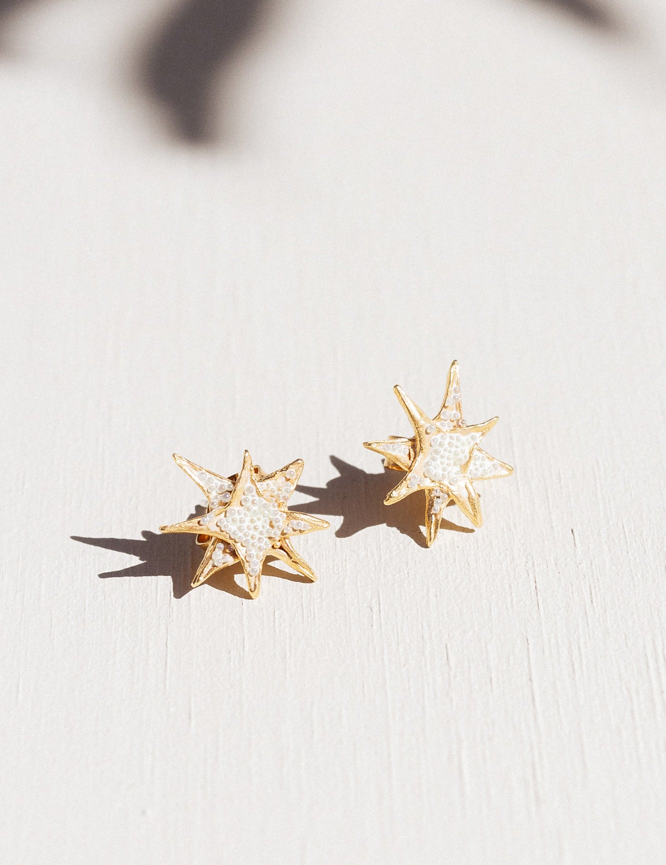 Dos aretes topos de estrella bañados en oro con detalles de cristales blancos, dispuestos sobre una superficie blanca con suaves sombras de hojas.
