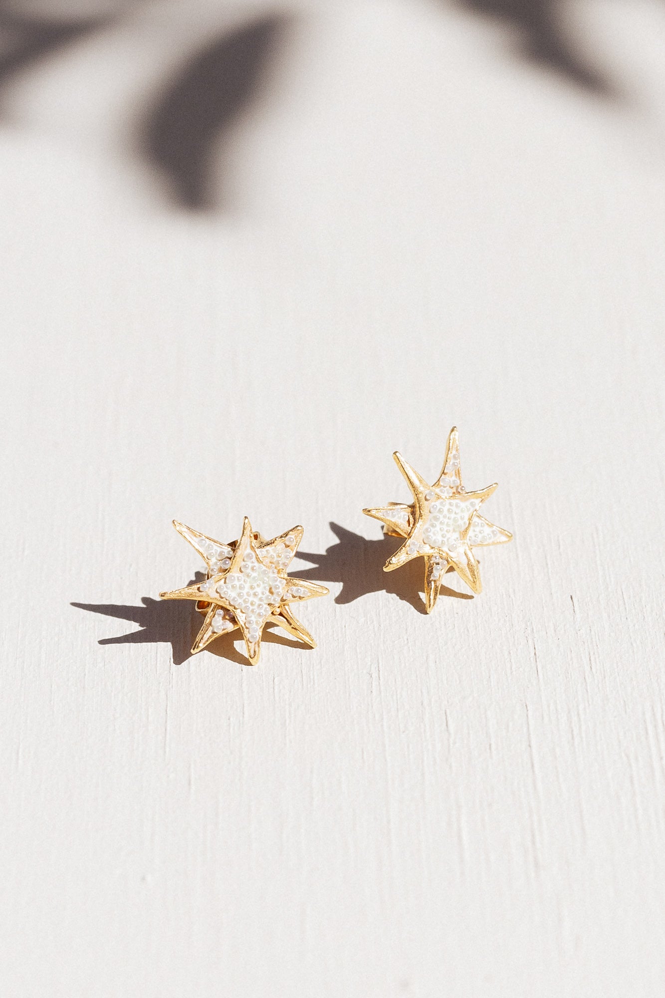 Dos aretes topos de estrella bañados en oro con detalles de cristales blancos, dispuestos sobre una superficie blanca con suaves sombras de hojas.