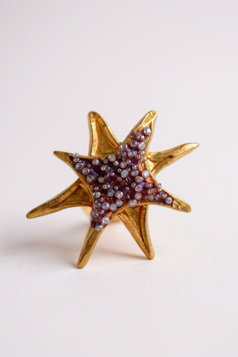 Un solo arete topos en forma de estrella bañados en oro con cristales púrpura, sobre fondo blanco limpio.