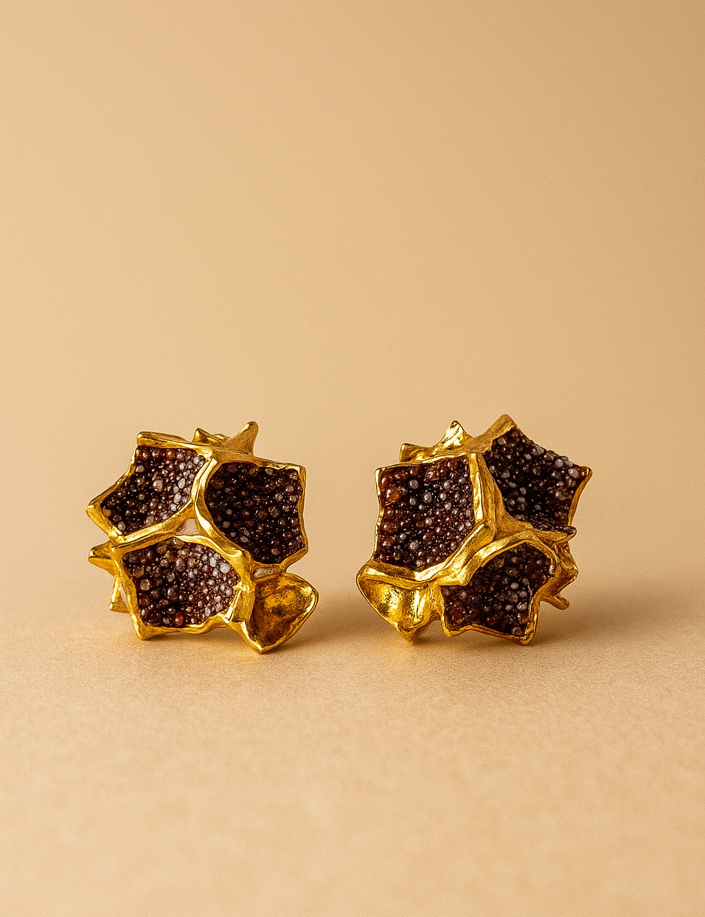 Primer plano de un par de aretes Galaxea en color rust: bañados en oro, con forma de estrella y cúmulos de gránulos de vidrio color óxido, sobre un fondo beige cálido.