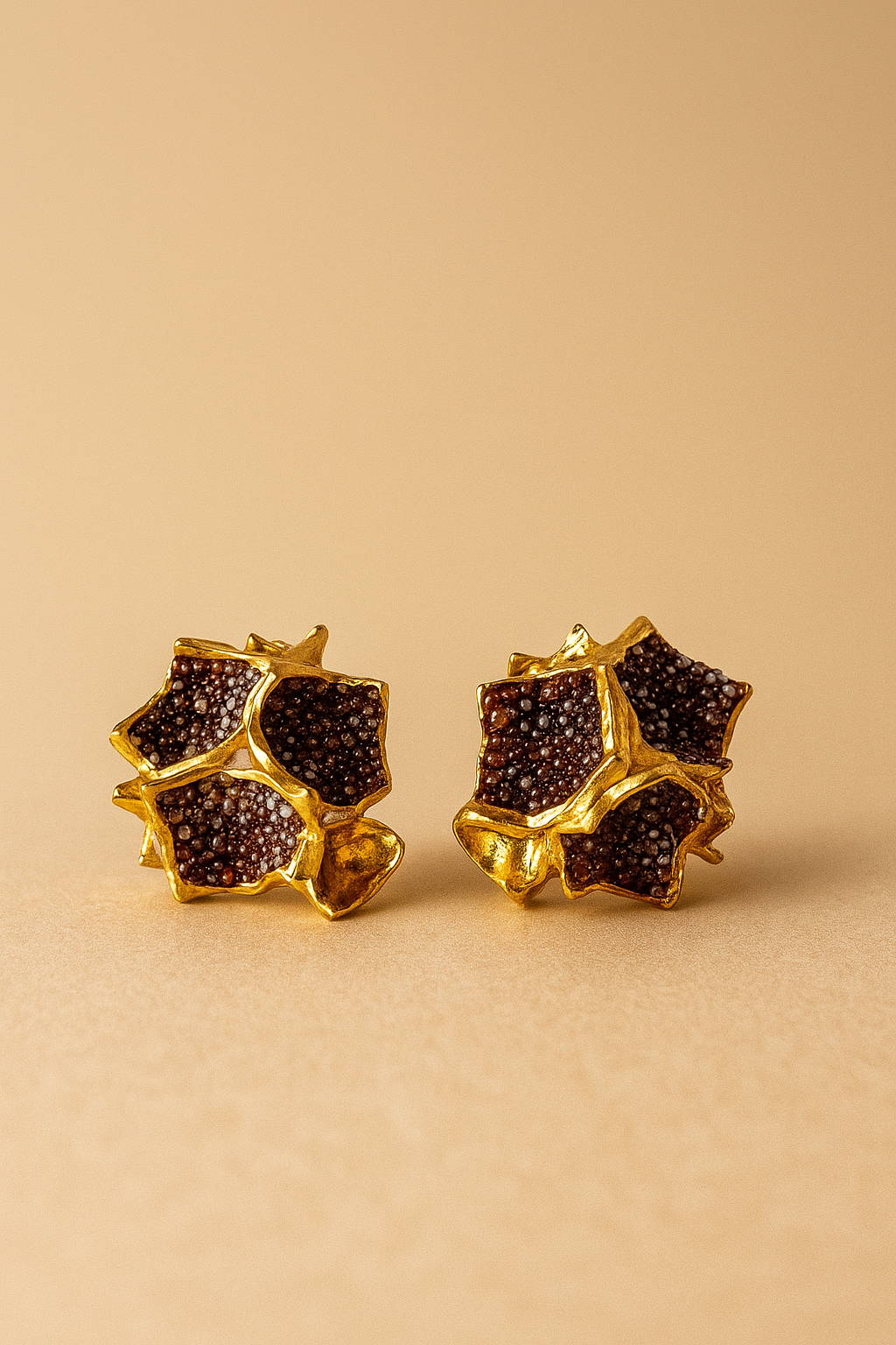 Primer plano de un par de aretes Galaxea en color rust: bañados en oro, con forma de estrella y cúmulos de gránulos de vidrio color óxido, sobre un fondo beige cálido.