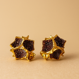 Primer plano de un par de aretes Galaxea en color rust: bañados en oro, con forma de estrella y cúmulos de gránulos de vidrio color óxido, sobre un fondo beige cálido.