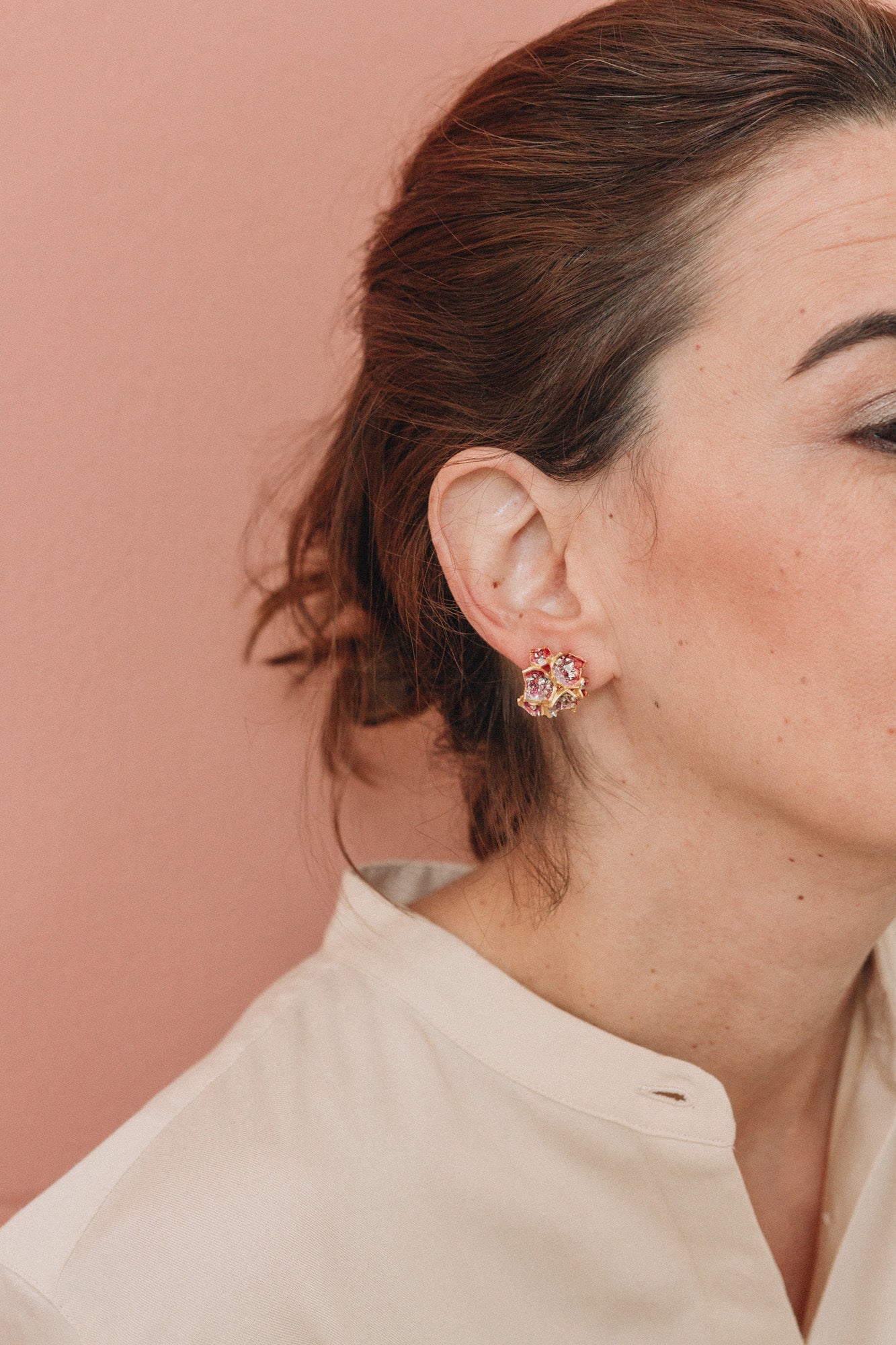 Perfil de una mujer luciendo un arete Galaxea en color Rust, bañado en oro con gránulos de vidrio, cabello recogido, sobre un fondo terracota suave.