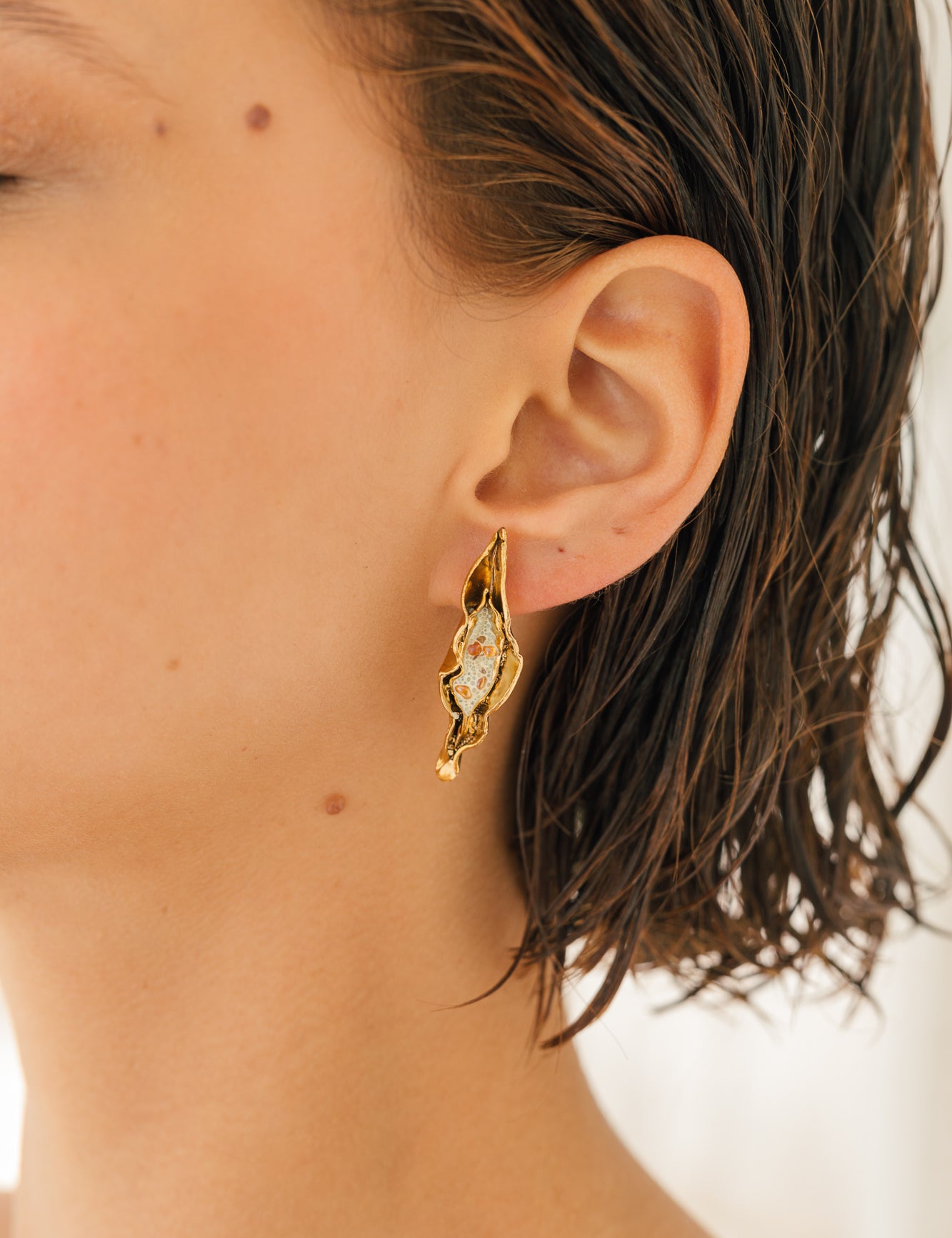 Primer plano lateral de la oreja de una mujer luciendo la variante Mostaza del Arete Lechuga, pintado a mano en amarillo mostaza con granulación de vidrio en montura de bronce bañado en oro, cabello recogido, sobre fondo neutro.