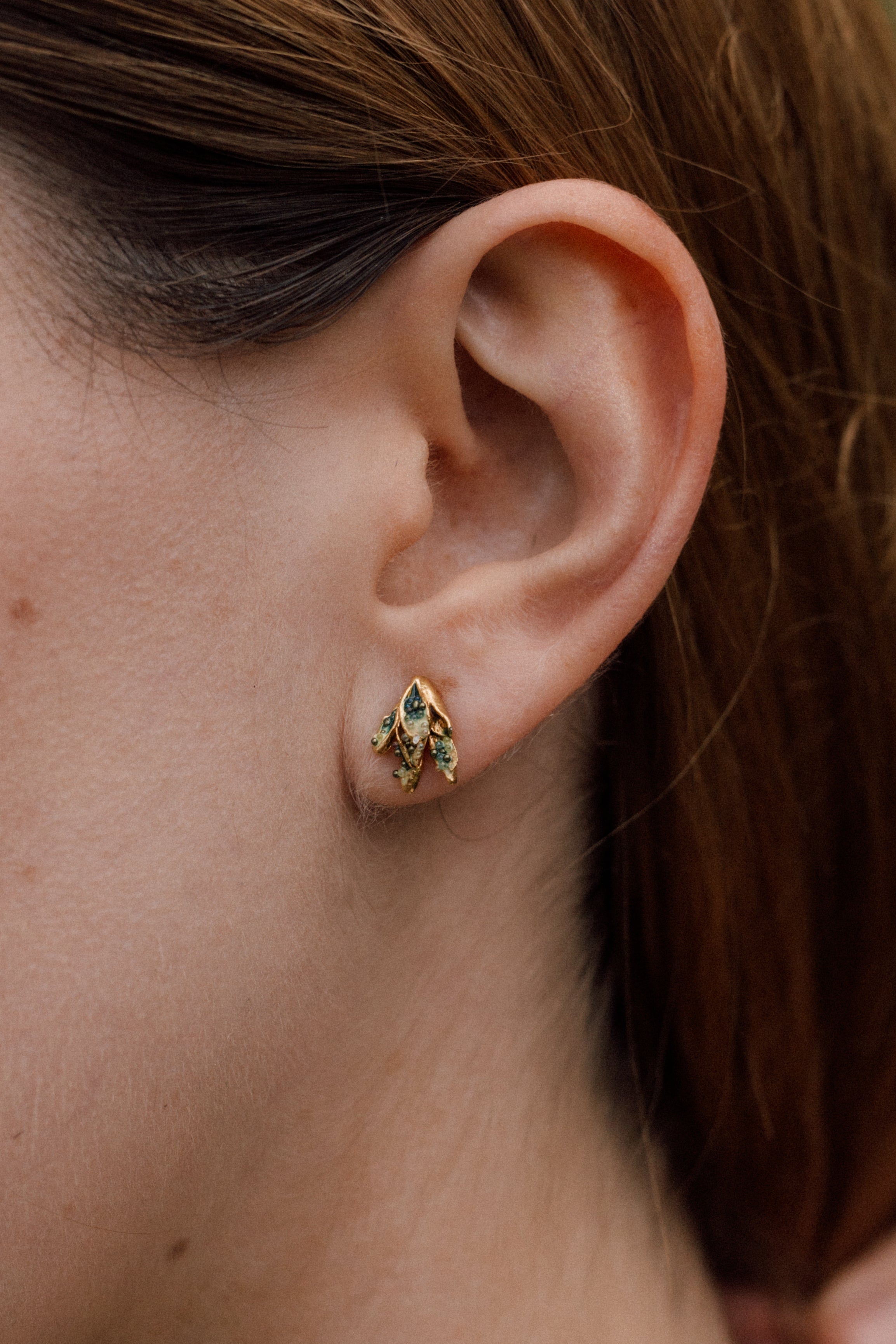 Primer plano lateral de la oreja de una mujer con arete topos Nebula Mini en bronce bañado en oro, pintado a mano en verde con granulación de vidrio, cabello recogido contra un fondo neutro.