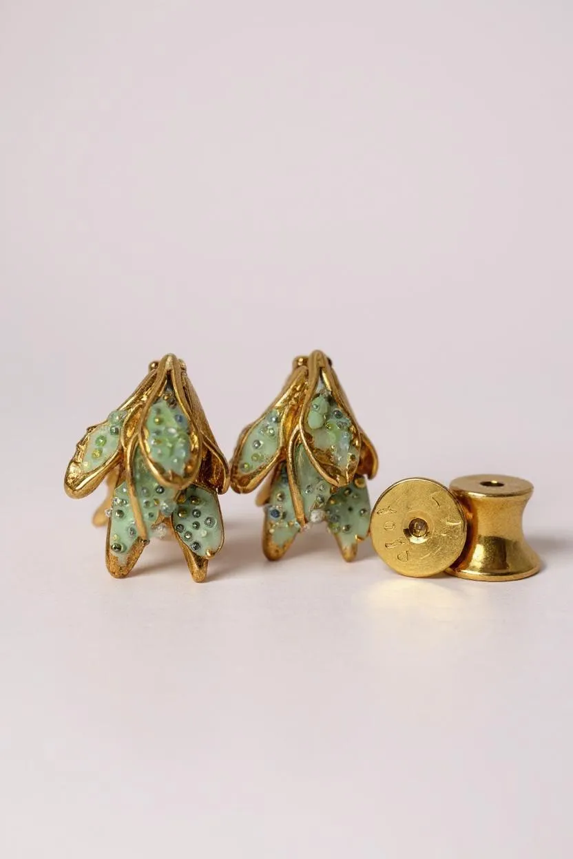Disposición plana de dos aretes Topos Nebula Mini en forma de hoja pintados a mano en verde con granulación de vidrio, acompañados de un topo redondo de bronce bañado en oro colocado aparte, sobre superficie blanca.