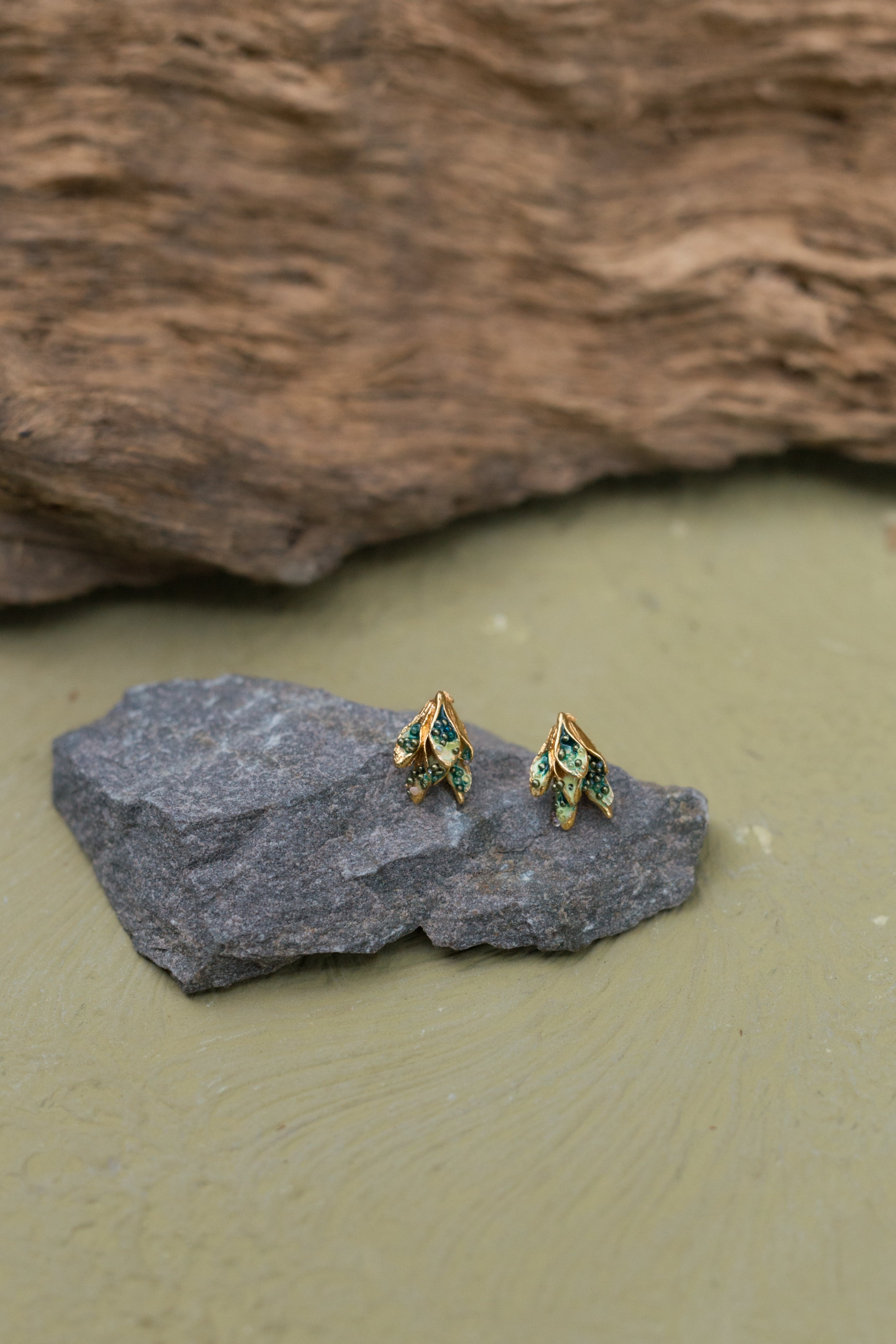 Dos topos Nebula Mini en forma de hoja con montura en bronce bañado en oro, pintados a mano en verde y con granulación de vidrio, reposando sobre una piedra gris sobre fondo oliva.