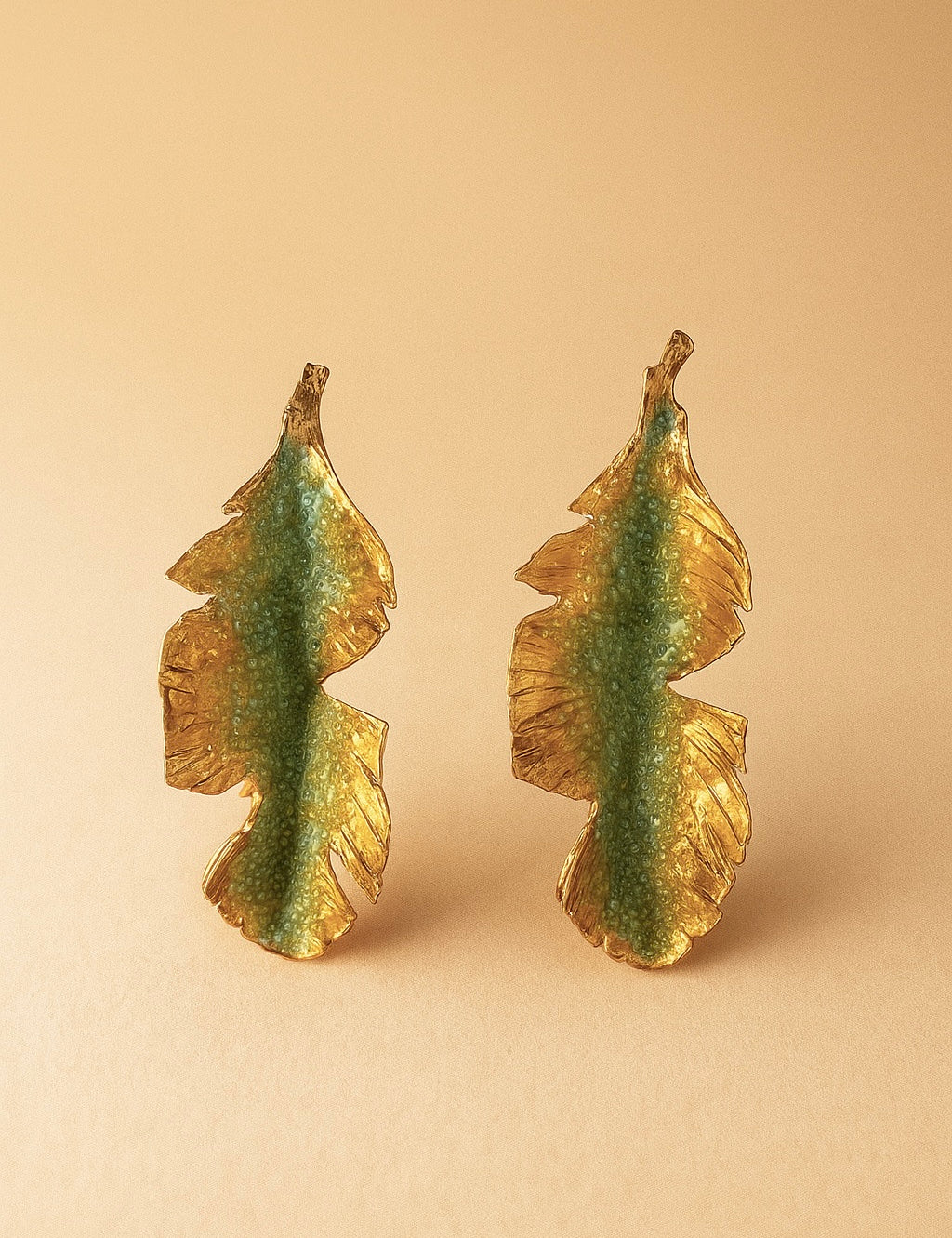Par de aretes Hojas de Plátano en bronce bañado en oro pintado a mano en verde sage detalle de granulación de vidrio, sobre fondo durazno suave.