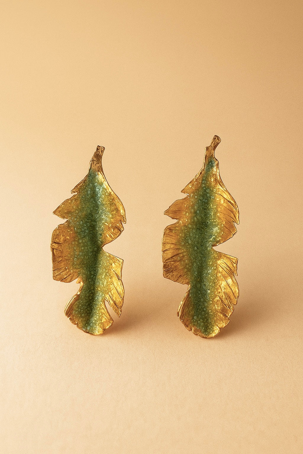Par de aretes Hojas de Plátano en bronce bañado en oro pintado a mano en verde sage detalle de granulación de vidrio, sobre fondo durazno suave.