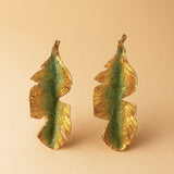 Par de aretes Hojas de Plátano en bronce bañado en oro pintado a mano en verde sage detalle de granulación de vidrio, sobre fondo durazno suave.