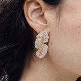 Primer plano lateral de la oreja de una mujer luciendo un arete Hojas de Plátano pintado en verde salvia con granulación de vidrio en montura de bronce bañado en oro, cabello recogido, sobre fondo neutro.