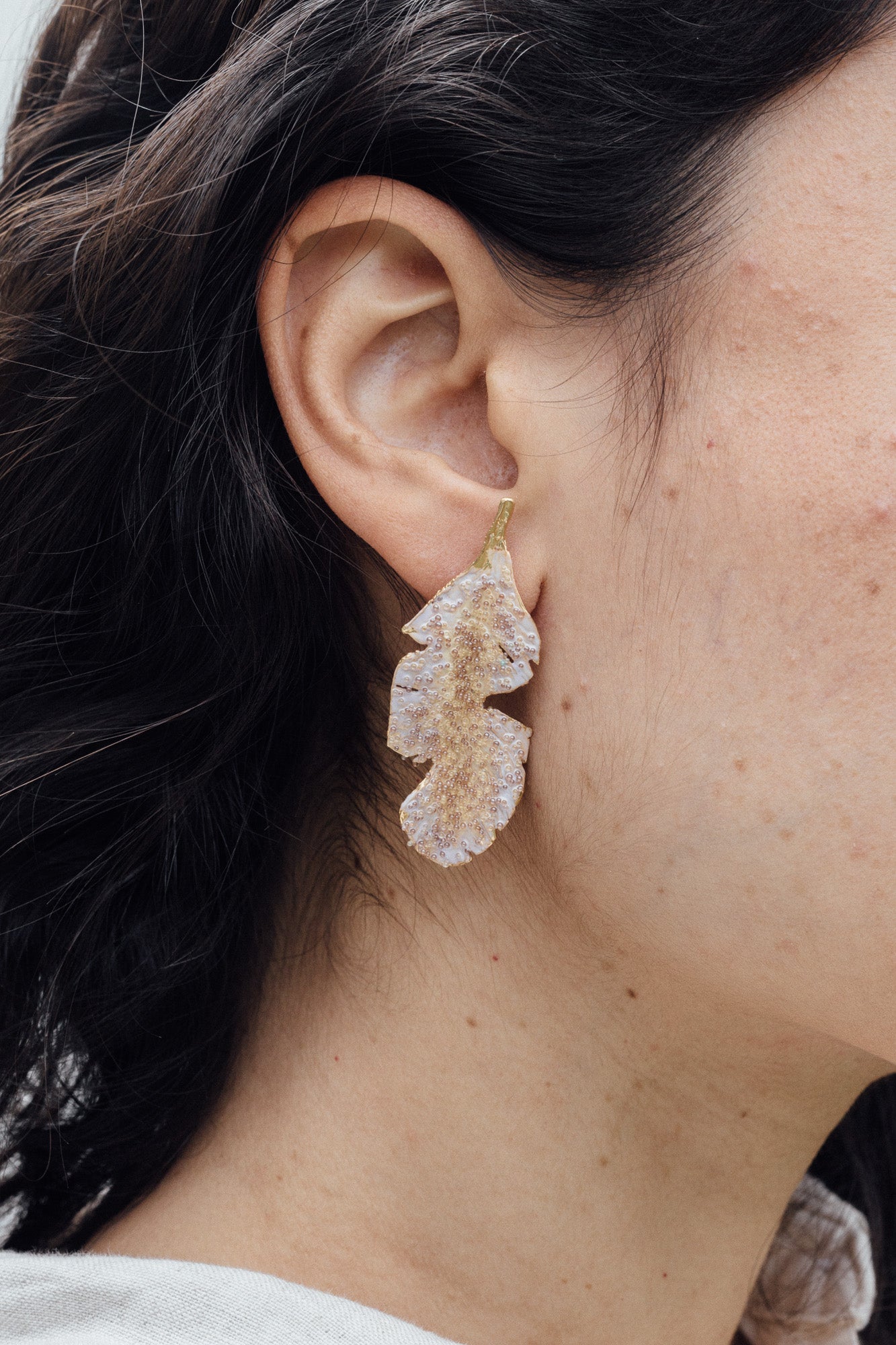 Primer plano lateral de la oreja de una mujer luciendo un arete Hojas de Plátano pintado en verde salvia con granulación de vidrio en montura de bronce bañado en oro, cabello recogido, sobre fondo neutro.