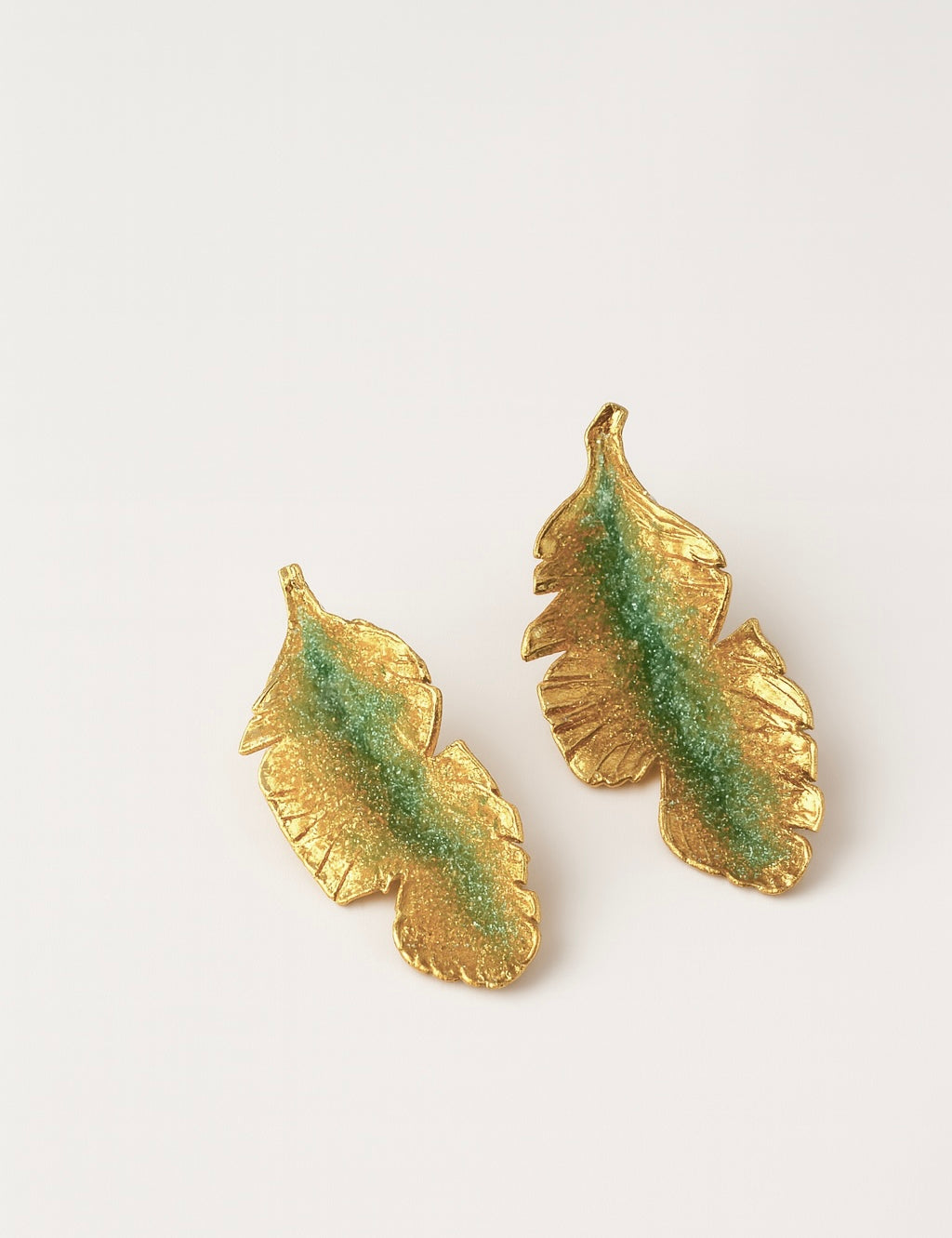 Par de aretes Hojas de Plátano en bronce bañado en oro pintado a mano en verde sage detalle de granulación de vidrio, sobre fondo blanco.