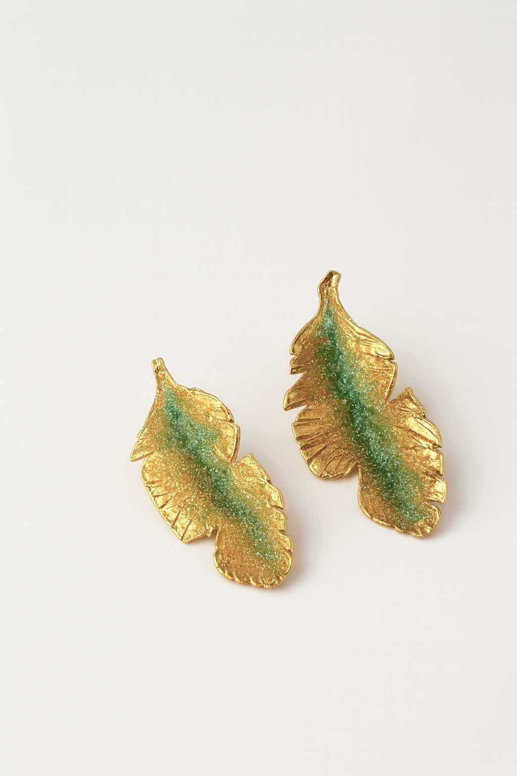 Par de aretes Hojas de Plátano en bronce bañado en oro pintado a mano en verde sage detalle de granulación de vidrio, sobre fondo blanco.