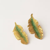 Par de aretes Hojas de Plátano en bronce bañado en oro pintado a mano en verde sage detalle de granulación de vidrio, sobre fondo blanco.