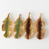 Disposición plana de cuatro aretes Hojas de Plátano alineados horizontalmente sobre fondo blanco—dos pintados a mano en verde salvia y dos en rust—con granulación de vidrio y montura de bronce bañado en oro.